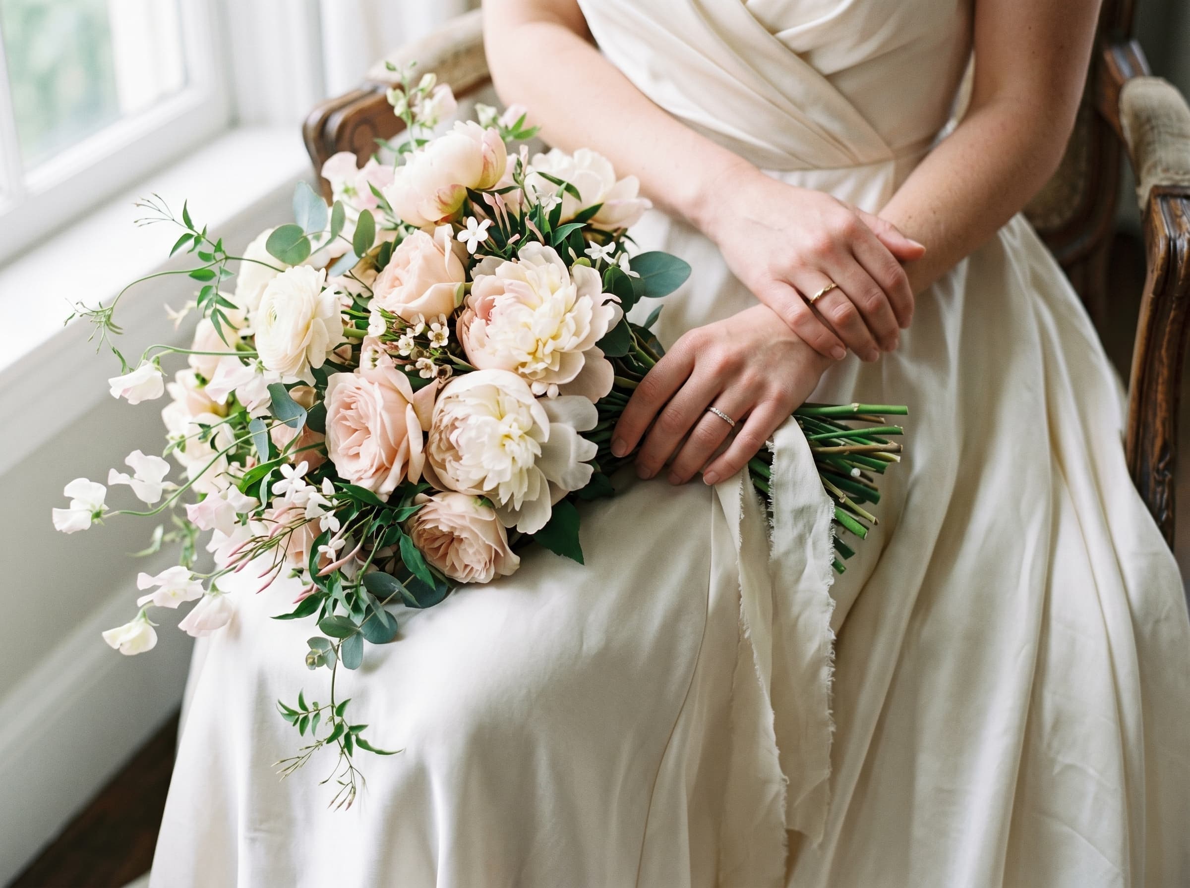 Bride's hands holding cream peonies and blush garden roses against a silk dress