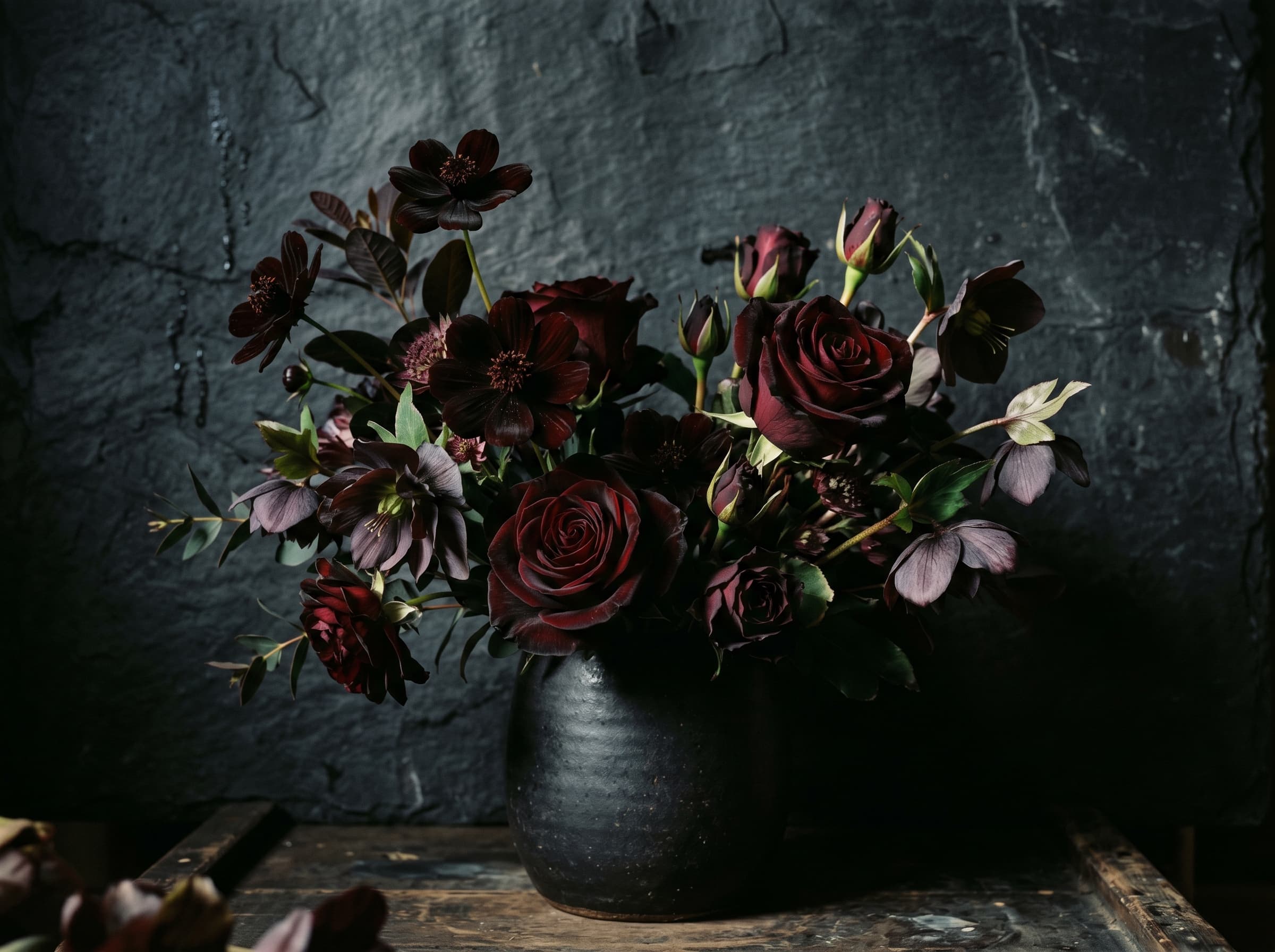 Moody arrangement of near-black dahlias and burgundy roses in a dark vessel against slate